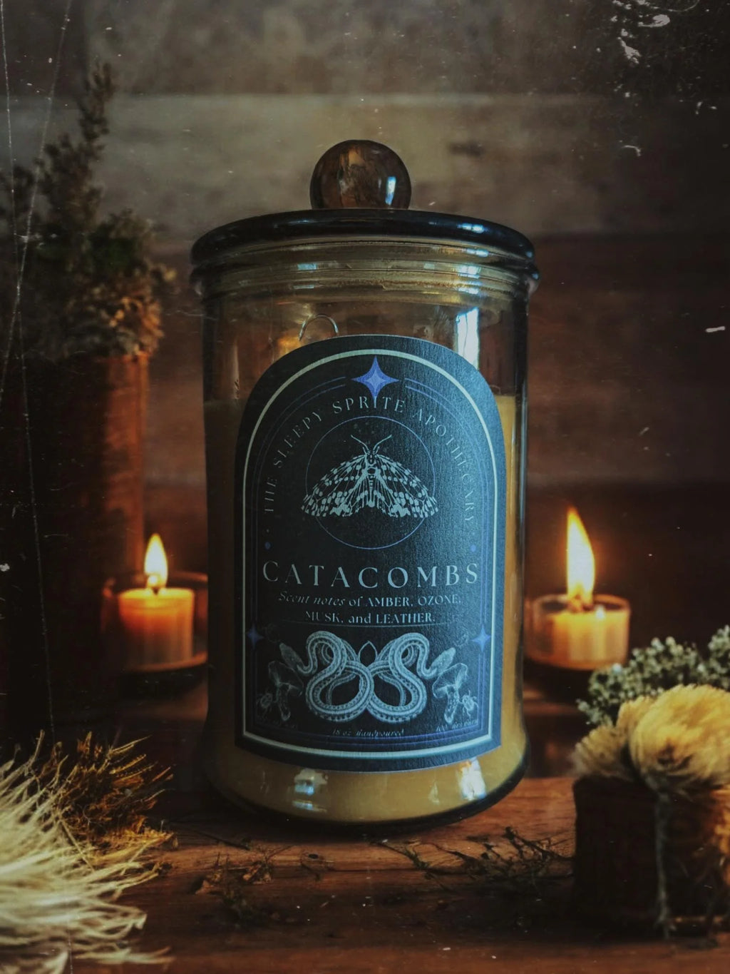 Candle jar labeled 'Catacombs' with decorative elements on a wooden surface. From the Sleepy Sprite Apothecary. Witchy dark cottagecore aesthetic.