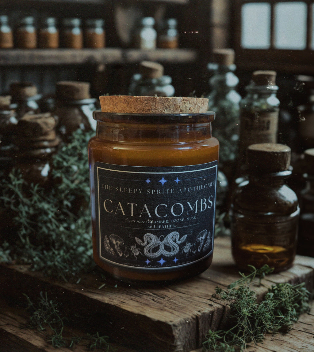 Candle jar labeled 'Catacombs' surrounded by other bottles and herbs on a wooden surface. From the Sleepy Sprite Apothecary. Witchy dark cottagecore aesthetic.