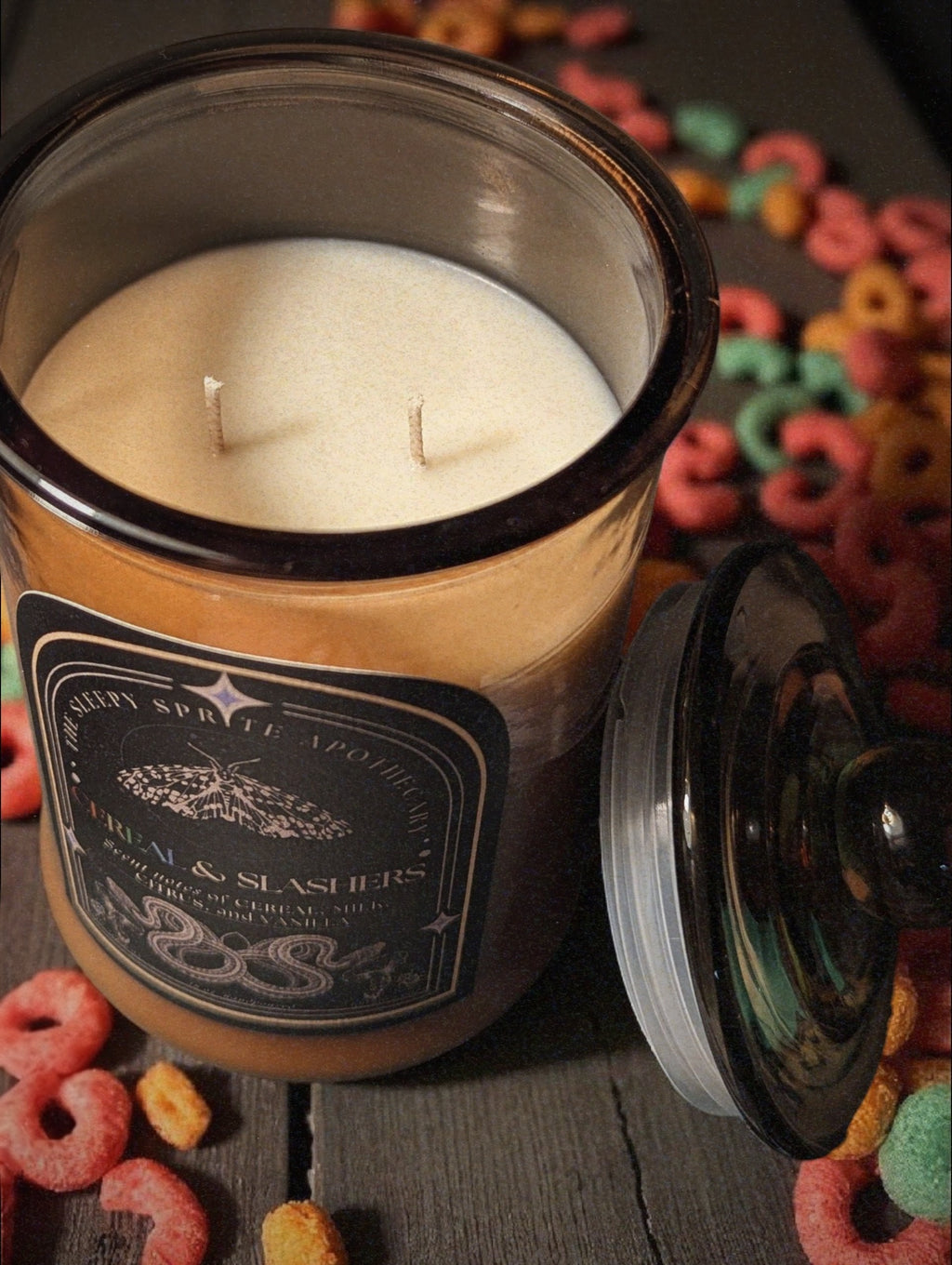Candle in a decorative jar with a label on a surface with colorful objects. Fruit loops scented candle. Witchy dark cottagecore aesthetic.