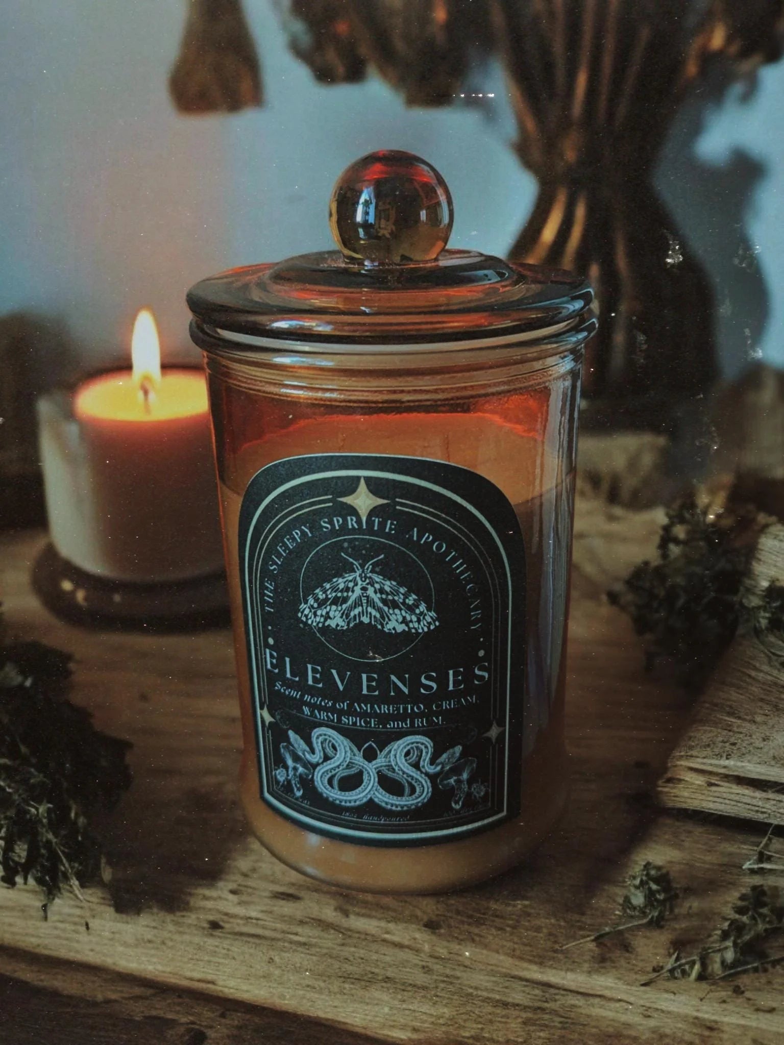Candle in a jar with a decorative label on a wooden surface, surrounded by candles and decorative items. Elevenses hobbit and Lord of the rings candle. Witchy dark cottagecore aesthetic.