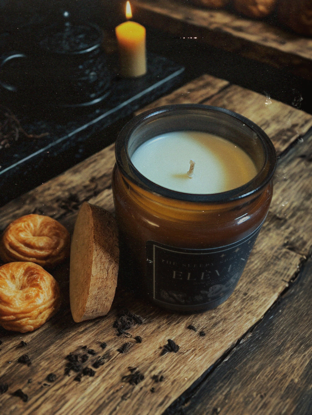 Elevenses Candle in a glass jar on a wooden surface with a lit candle and pastries in the background. Hobbit and Lord of the rings candle.Witchy dark cottagecore aesthetic.