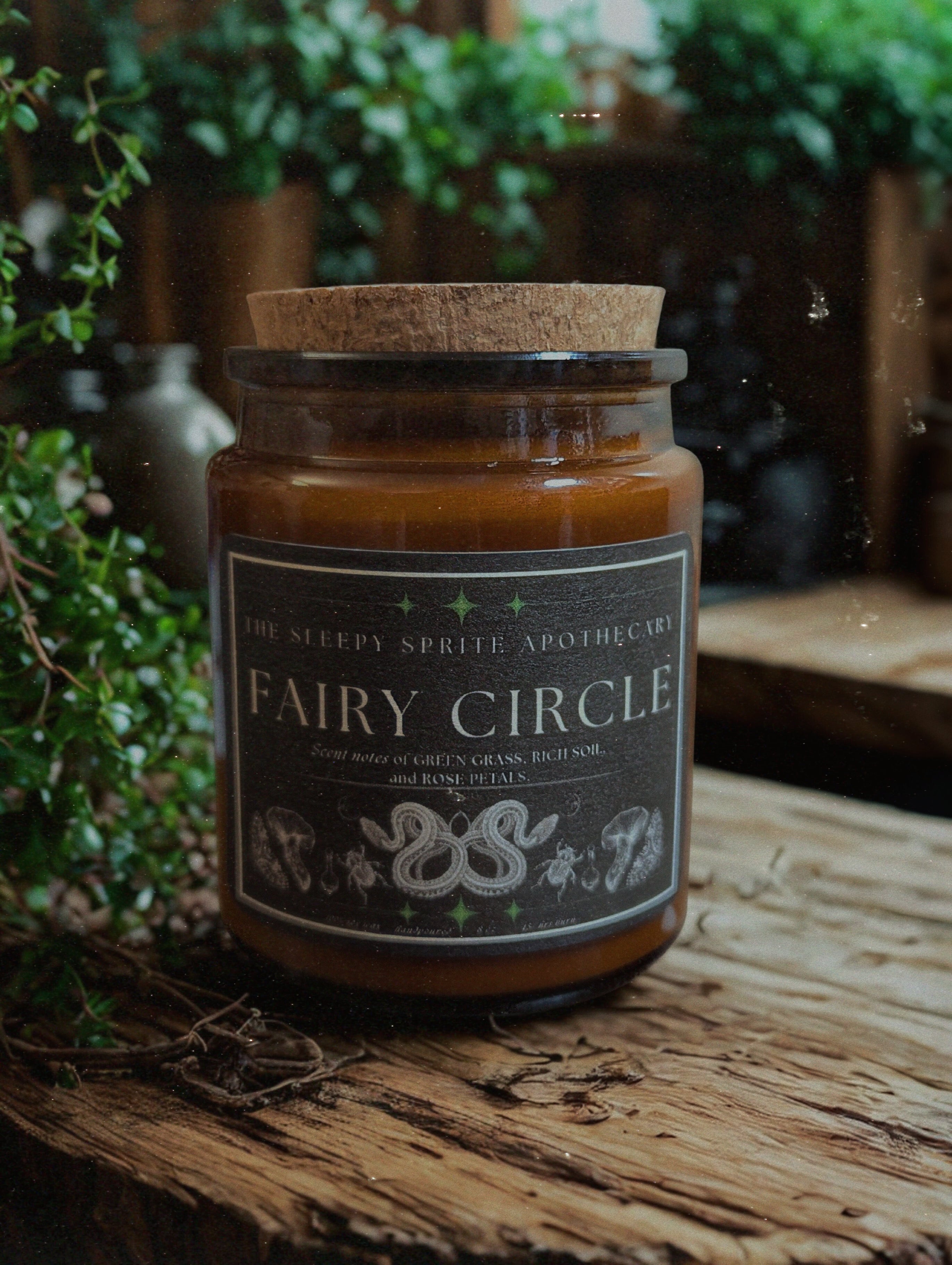 Candle jar labeled 'Fairy Circle' on a wooden surface with greenery in the background from The Sleepy Sprite Apothecary. Witchy dark cottagecore aesthetic.