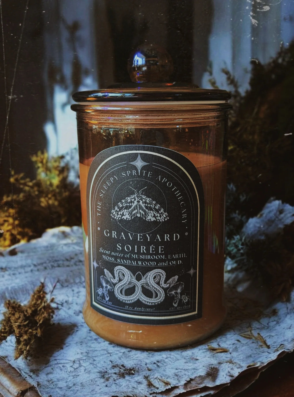 Candle jar labeled 'Graveyard Soireé' with a dark label against a natural background from The Sleepy Sprite Apothecary. Witchy dark cottagecore aesthetic.