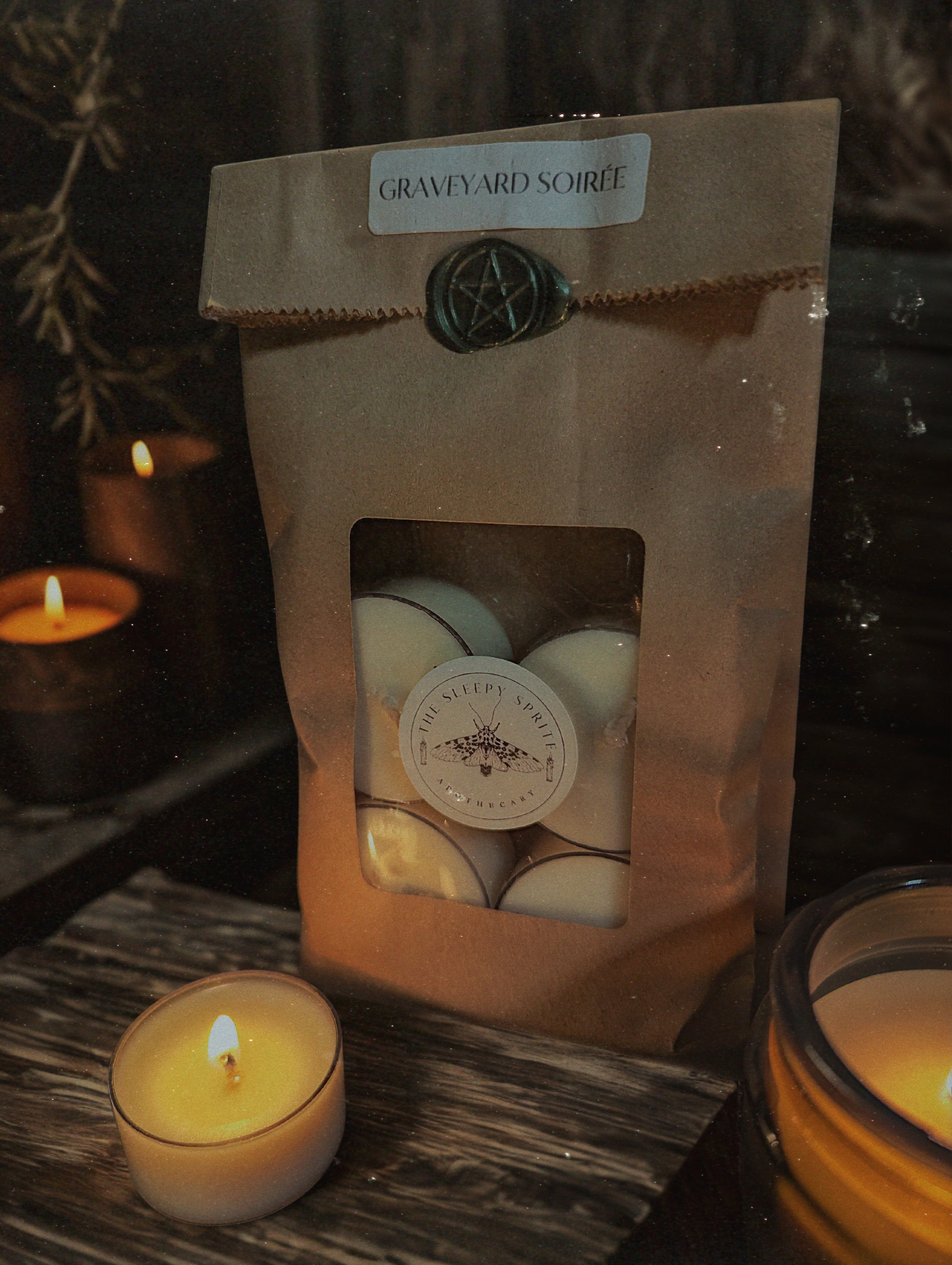 Packaged candles with a lit candle in front on a wooden surface from the Sleepy Sprite Apothecary.Witchy dark cottagecore aesthetic.