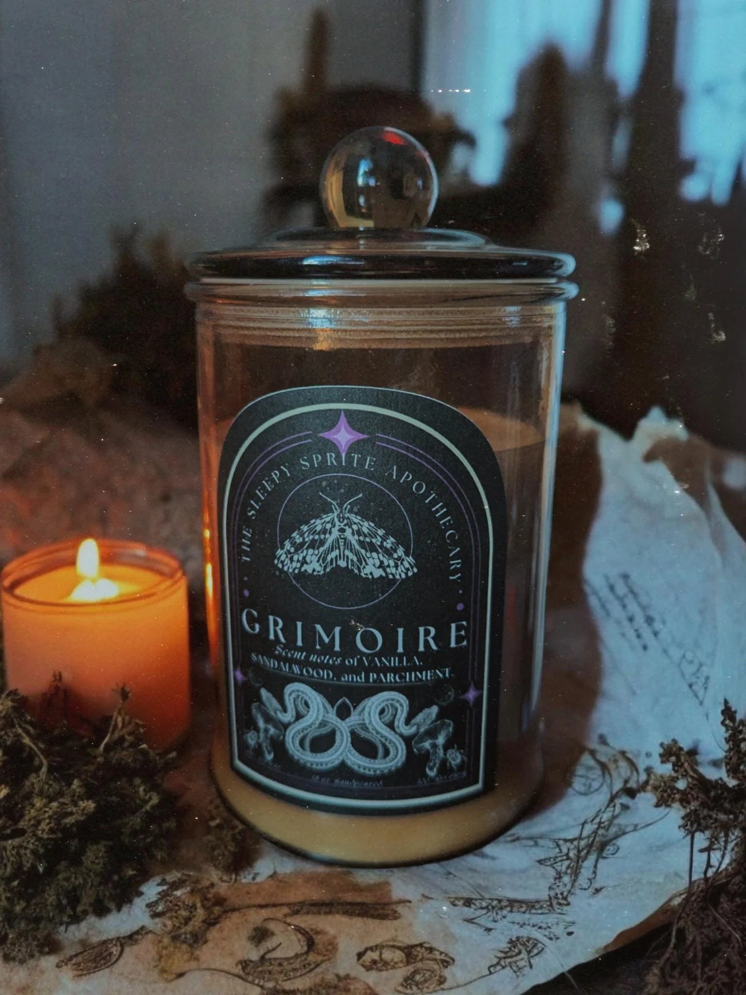 Glass jar labeled 'Grimoire' with a candle in the background from The Sleepy Sprite Apothecary. Witchy dark cottagecore aesthetic.