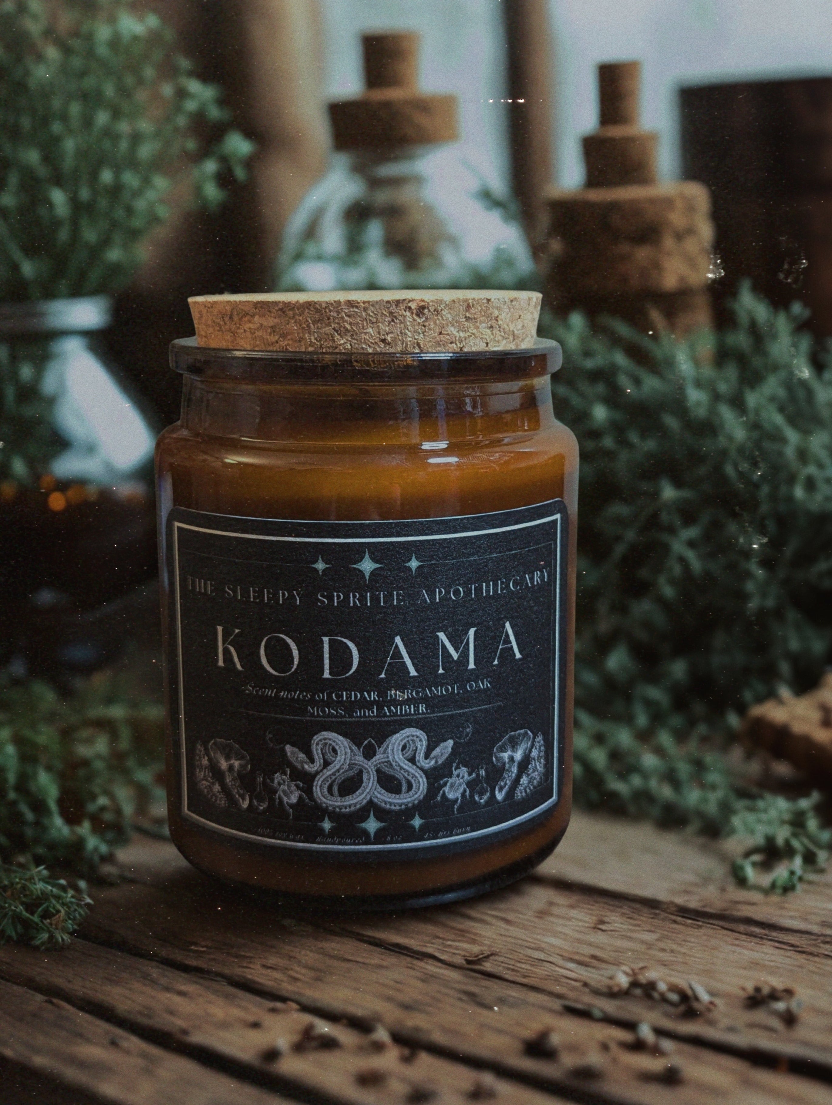 Jar of 'Kodama' candle with cork lid on a wooden surface surrounded by greenery from The Sleepy Sprite Apothecary. Witchy dark cottagecore aesthetic.