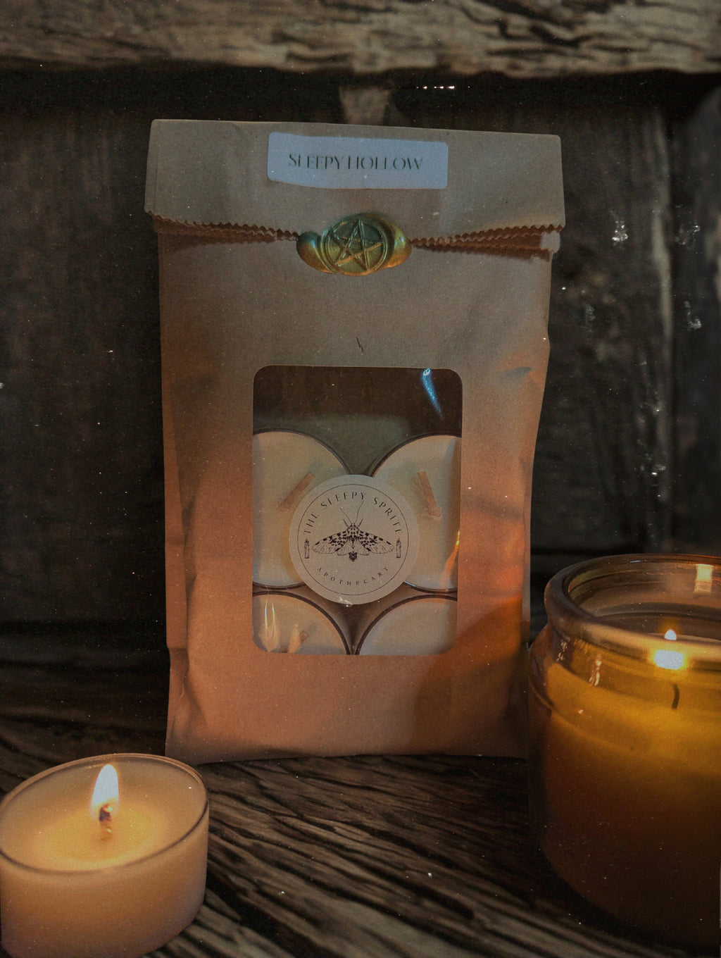 Packaged candle with 'Sleepy Hollow' branding from The Sleepy Sprite Apothecary next to a lit candle on a wooden surface. Witchy dark cottagecore aesthetic.