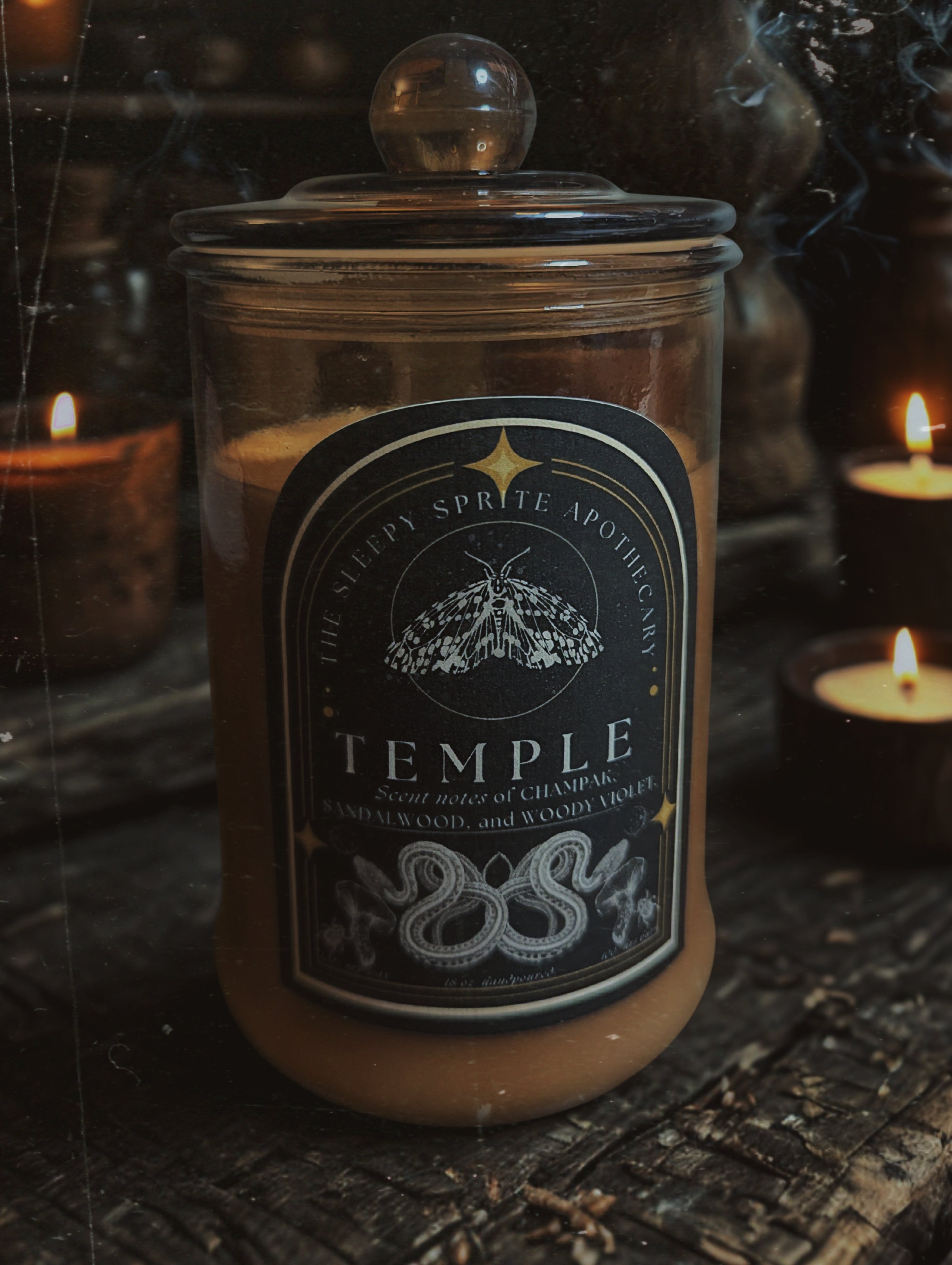 Candle jar labeled 'Temple' from 'The Sleepy Sprite Apothecary' with candles in the background. Witchy dark cottagecore aesthetic.