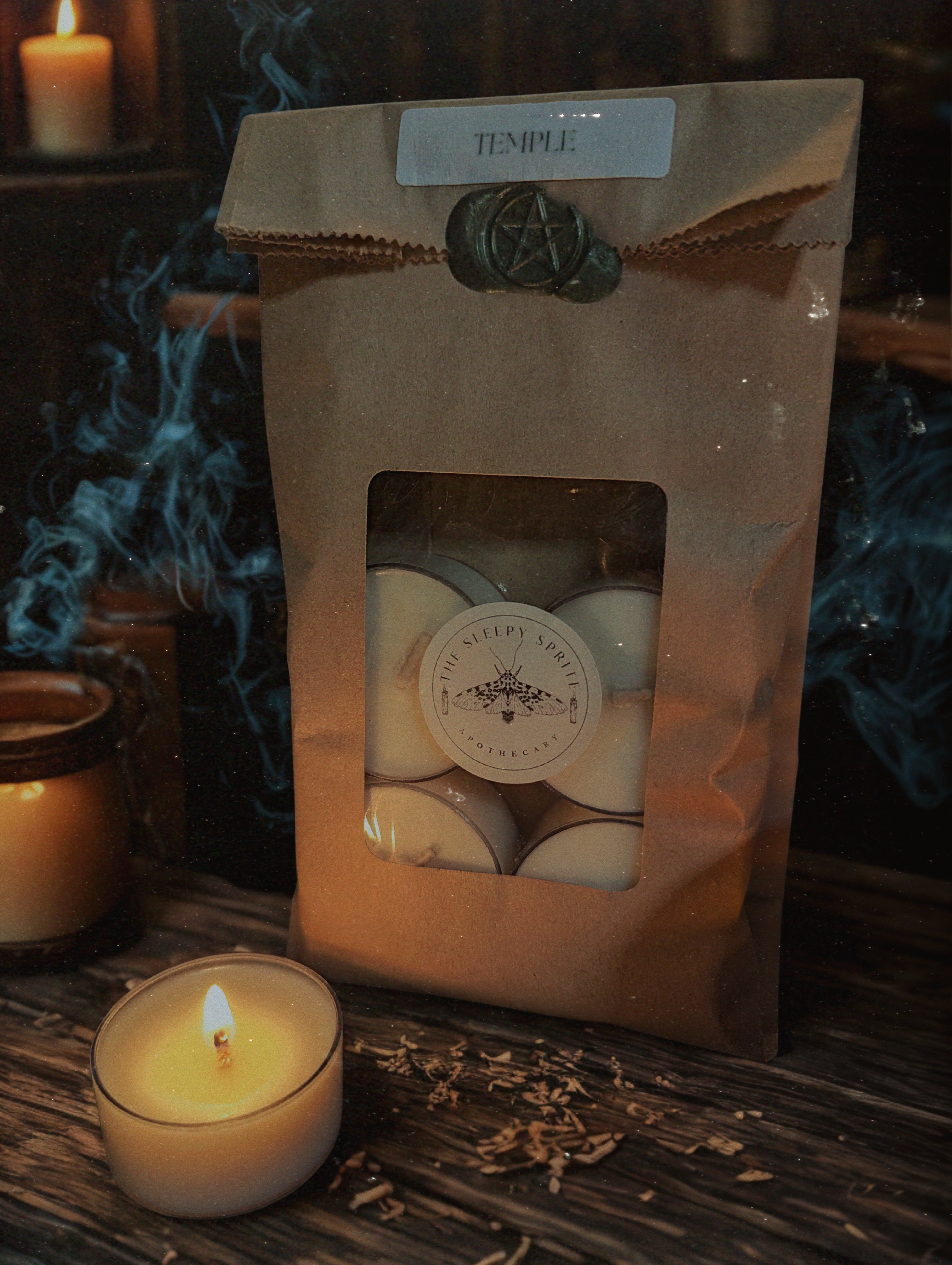 Packaged tea light  candles from the Sleepy Sprite Apothecary on a dark background with a lit candle. Witchy dark cottagecore aesthetic.