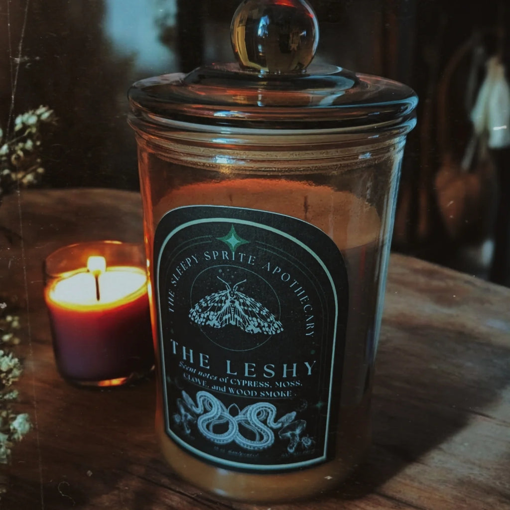 Leshy soy Candle in a glass jar with a label on a wooden surface, surrounded by other candles. Witchy dark cottagecore aesthetic.