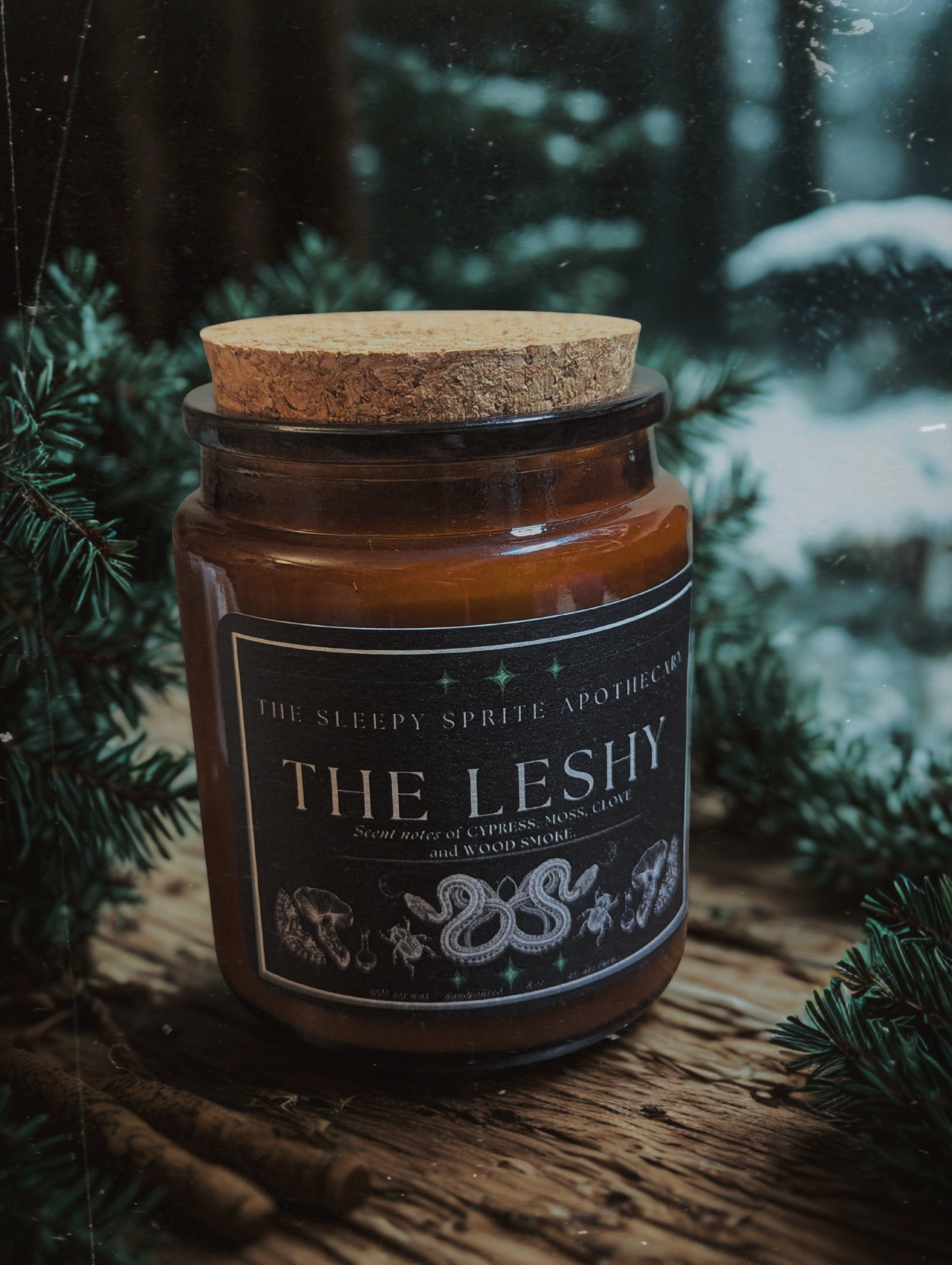 Candle jar labeled 'The Leshy' with a cork lid on a wooden surface with a forest background from The Sleepy Sprite Apothecary. Witchy dark cottagecore aesthetic.