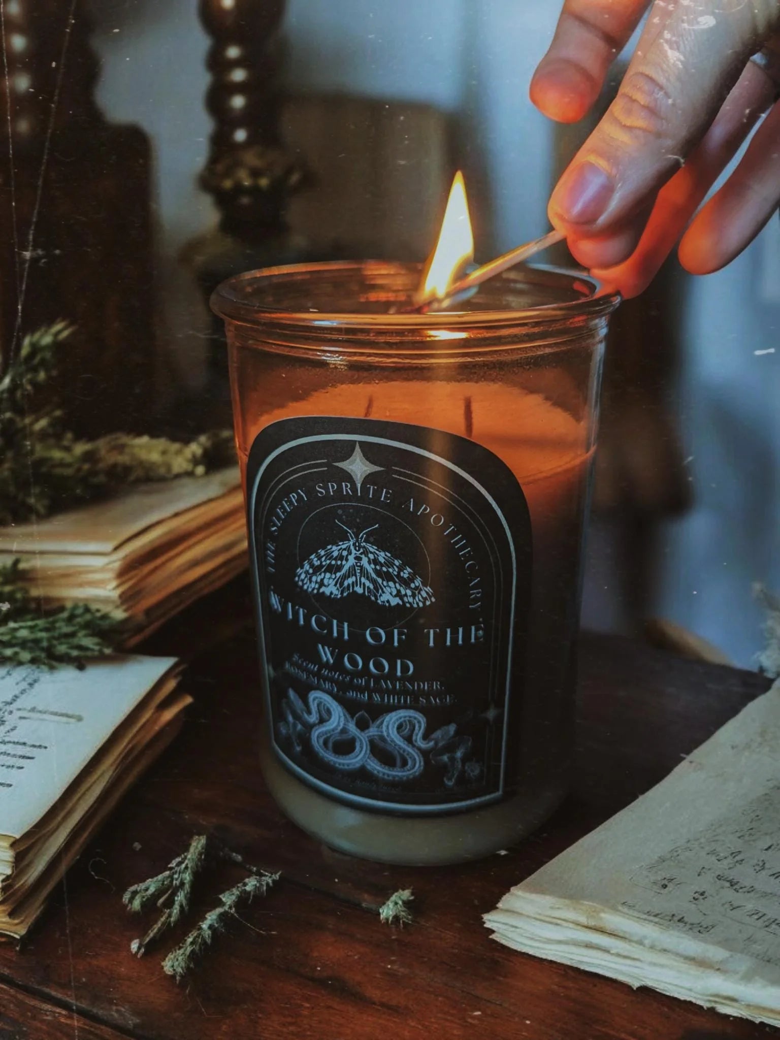 Witch of the woods Candle being lit with a match, surrounded by books and herbs on a wooden surface. Witchy dark cottagecore aesthetic. Baba yaga inspired.